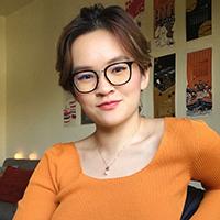 A person wearing glasses and an orange shirt, seated in front of a wall with posters