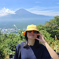 A person wearing a blue shirt, yellow hat, and sunglasses, posing in front of a scenic mountain overlook