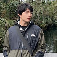 A person in an olive and black raincoat standing on a bridge over flowing water running through a tropical grove