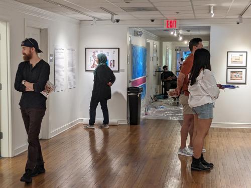 people standing in an art gallery looking at pictures; the artist (Venazir Martinez) speaks with one visitor