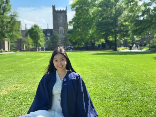 A person wearing graduation regalia sitting on a grassy lawn on Duke's campus