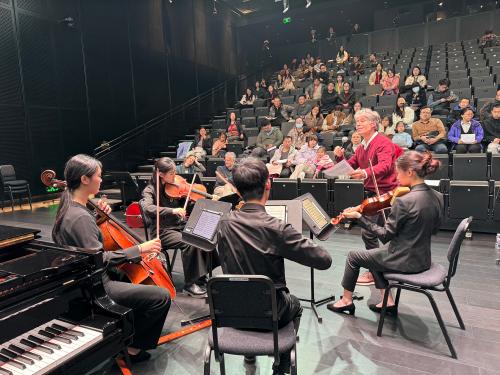 Stage-level view of people playing instruments on a stage while spectators sit in the audience section of a theater