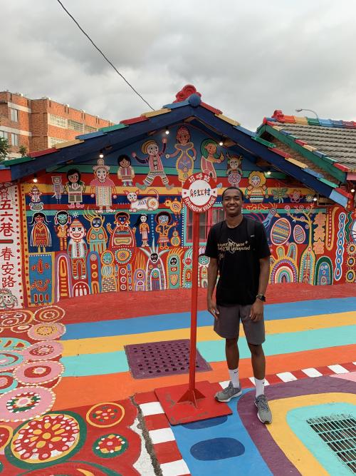 A person standing by a signpost in front of a brightly-painted building and roadway